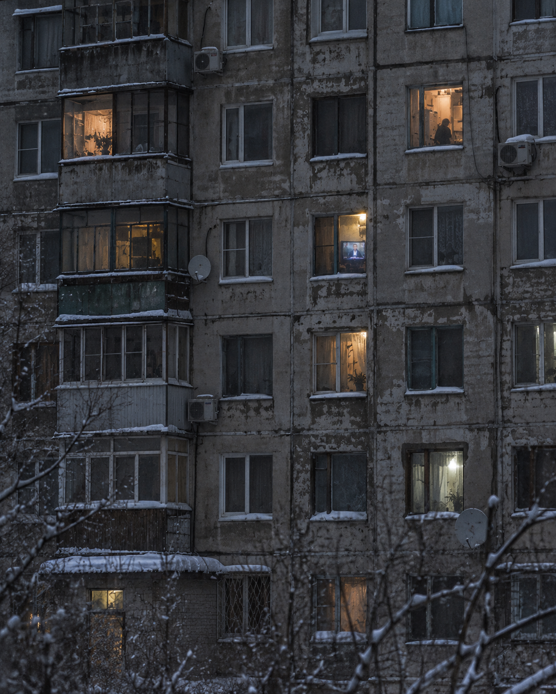 Weathered winter apartment block with scattered warm windows glowing against snow